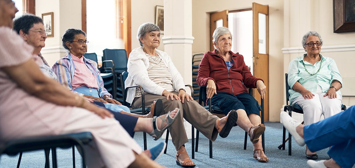 Group of senior women in assisted living facility participate in a seated exercise class.